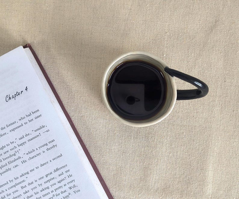 May include: A cup of dark coffee with a black handle sits next to an open book on a beige linen surface. The book is open to Chapter 4. The coffee cup is white with a black handle.