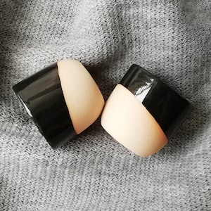 Może przedstawiać: Dwa ceramiczne kubki w kolorze czarnym i beżowym. Kubki są w połowie czarne, a w połowie beżowe, przy czym czarna część znajduje się na g&oacute;rze. Kubki stoją na szarej powierzchni z tkaniny.