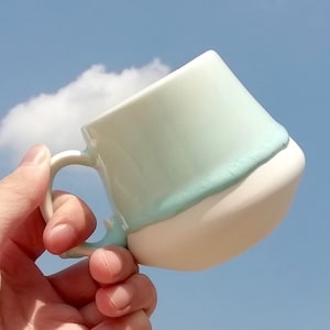 Puede incluir: Una taza de cerámica azul claro y blanco con un asa redondeada. La taza se sostiene en una mano contra un fondo de cielo azul.
