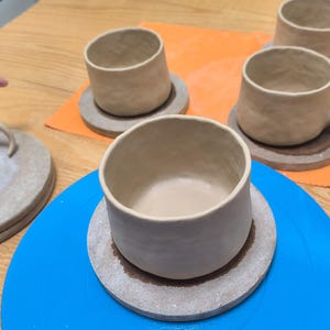 May include: Handmade ceramic mugs and coasters in various stages of completion. The mugs are a light beige color, with a textured surface. One mug has a handle. The coasters are round and a similar color. The items are on a blue and orange surface.