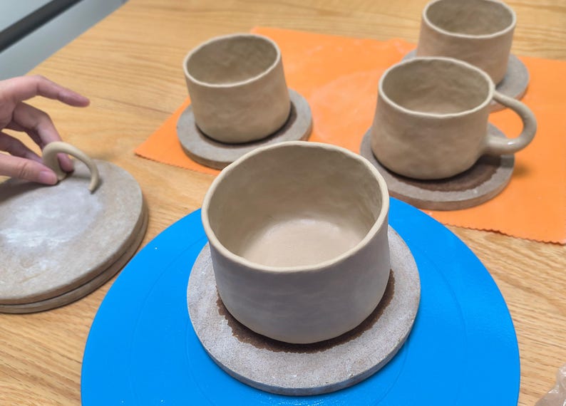 May include: Handmade ceramic mugs and coasters in various stages of completion. The mugs are a light beige color, with a textured surface. One mug has a handle. The coasters are round and a similar color. The items are on a blue and orange surface.