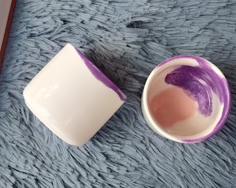 May include: Two ceramic cups with a white base and a purple and pink marbled design. The cups are on a blue fuzzy surface.