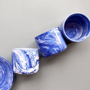 May include: Four blue and white marbled ceramic cups with a swirling pattern. The cups are arranged in a row on a gray background.