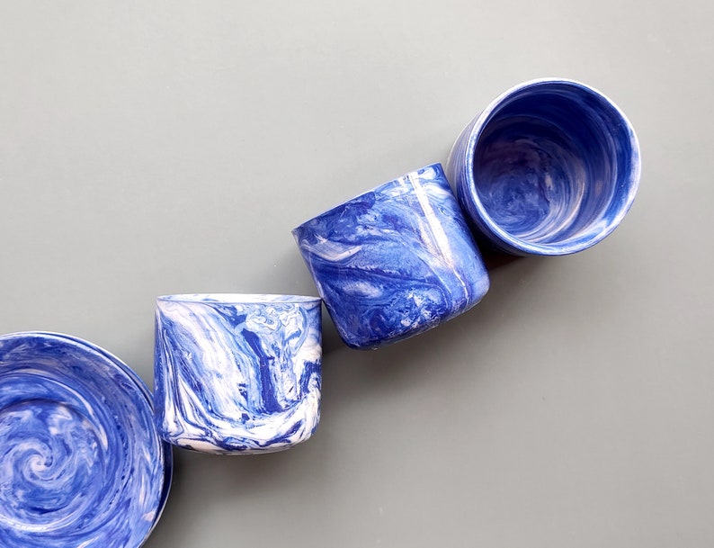 May include: Four blue and white marbled ceramic cups with a swirling pattern. The cups are arranged in a row on a gray background.