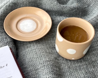 Puede incluir: Una taza de cerámica beige con lunares blancos llena de café, y un platillo beige a juego.