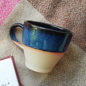 May include: A blue and white ceramic mug with a rounded handle. The mug has a dark blue glaze on the top half and a light cream glaze on the bottom half.