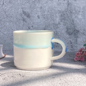 May include: White ceramic mug with a light blue glaze and a blue handle. The mug is sitting on a gray concrete surface.