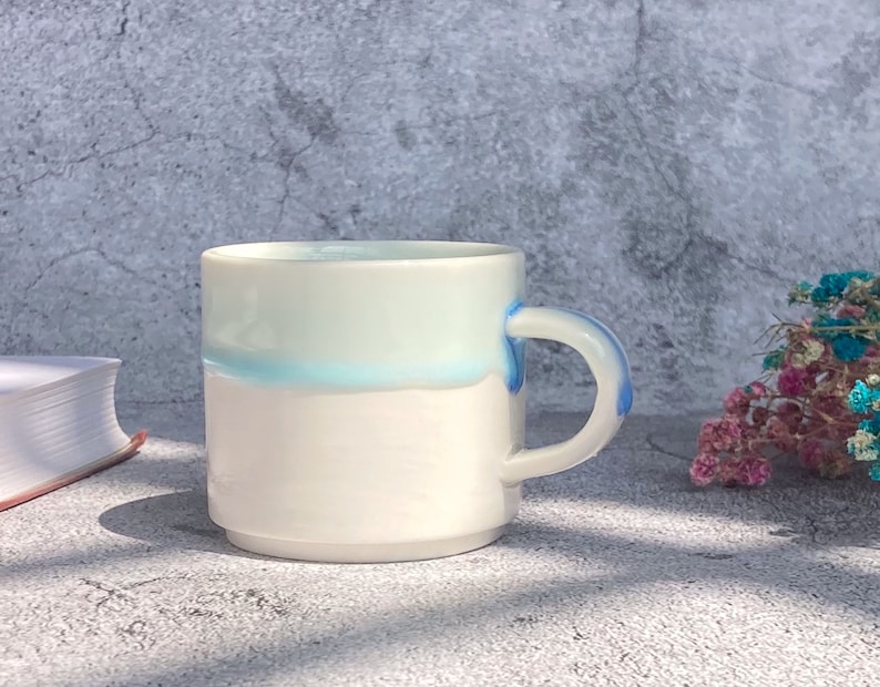 May include: White ceramic mug with a light blue glaze and a blue handle. The mug is sitting on a gray concrete surface.