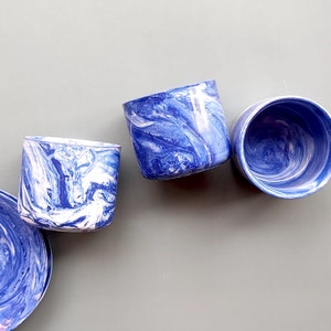 May include: Three blue and white marbled ceramic cups with a swirling pattern. The cups are arranged in a row on a light gray surface.