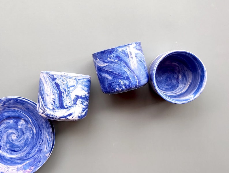 May include: Three blue and white marbled ceramic cups with a swirling pattern. The cups are arranged in a row on a light gray surface.