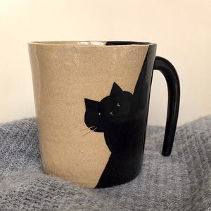 Puede incluir: Una taza de cerámica con un diseño de gato negro. La taza tiene un exterior beige con una ilustración de gato negro en un lado y un asa negra. El gato está pintado de negro y parece estar mirando por encima del borde de la taza.