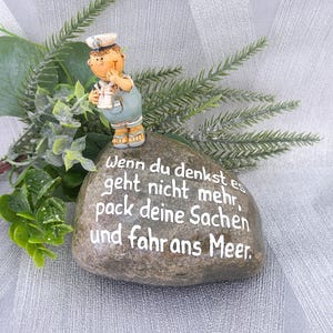 Puede incluir: Una piedra decorativa con el texto alemán "Wenn du denkst es geht nicht mehr, pack deine Sachen und fahr ans Meer." Una pequeña figura de marinero está encima. Hay vegetación en el fondo.