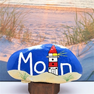 Puede incluir: Piedra pintada a mano con fondo azul, arena blanca y un faro rojo y blanco con techo amarillo. La palabra "Moin" está escrita en blanco en la piedra.