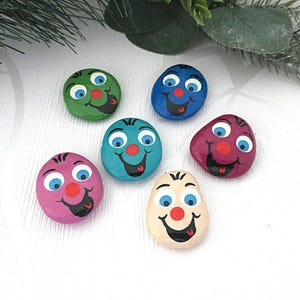Può includere: Sei sassi dipinti con facce sorridenti in vari colori: verde, blu, verde acqua, rosa, viola e beige. Ogni sasso presenta occhi blu, un naso rosso e una bocca sorridente. Arte decorativa.