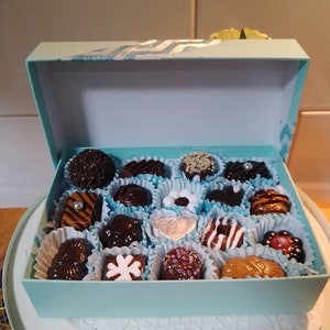 May include: A light blue rectangular gift box filled with an assortment of sixteen individual chocolates. The chocolates vary in shape, colour, and topping, including dark, milk, and white chocolate pieces with sprinkles, drizzles, and foil.