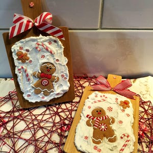 May include: Two wooden boards with gingerbread man decorations. Each board has a gingerbread man with a red and white scarf, candy canes, and red and white bows. The gingerbread men are on a white frosting-like base.