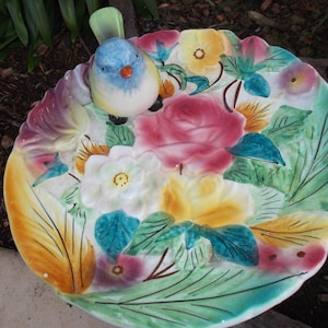 May include: A ceramic bird bath with a colourful floral design. The bird is blue, white, and yellow with an orange beak. The bath features pink, yellow, and white flowers with green leaves. The rim is scalloped.