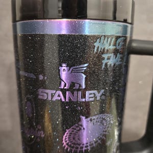 May include: Black Stanley travel mug with a handle, featuring a glittery finish and purple accents. The mug has the Stanley logo and the words "HALL OF FAME" printed on it. The design includes a winged creature and other graphics.