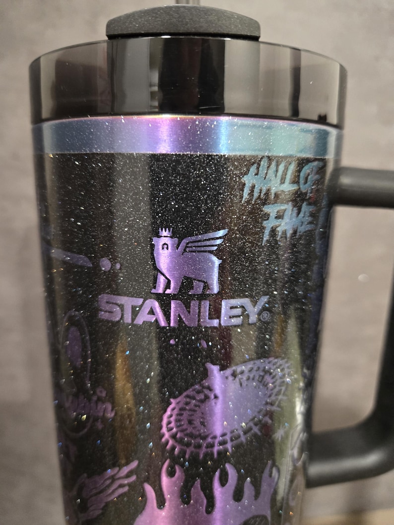 May include: Black Stanley travel mug with a handle, featuring a glittery finish and purple accents. The mug has the Stanley logo and the words "HALL OF FAME" printed on it. The design includes a winged creature and other graphics.