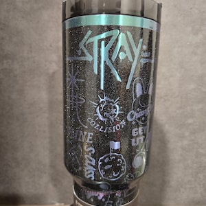 May include: A black glitter tumbler with teal accents and the word "STRAY". The tumbler is decorated with illustrations and text, including "COLLISION" and a cartoon rabbit. A clear straw is inserted into the lid.