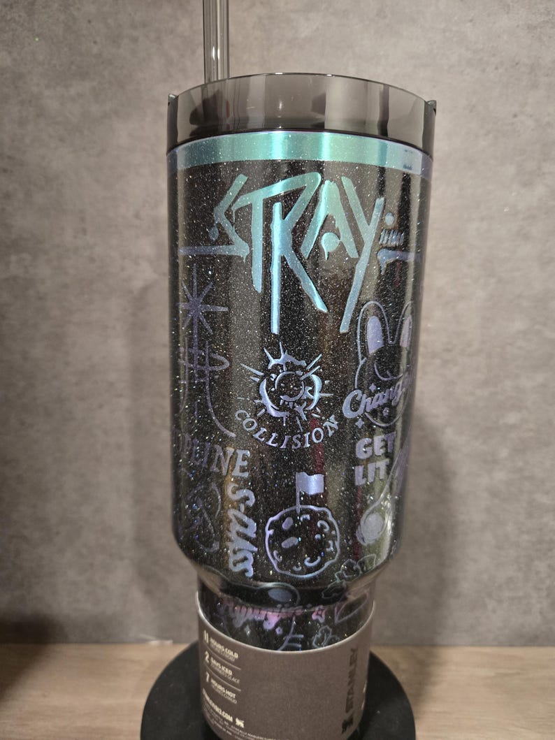 May include: A black glitter tumbler with teal accents and the word "STRAY". The tumbler is decorated with illustrations and text, including "COLLISION" and a cartoon rabbit. A clear straw is inserted into the lid.