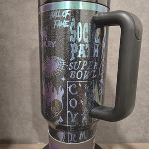 May include: Black glitter tumbler with a black handle and lid. The tumbler features holographic text and graphics, including "Hall of Fame," "Super Bowl," and "Social Path." The tumbler has a straw and is labeled "Quencher H2.0 Tumbler."