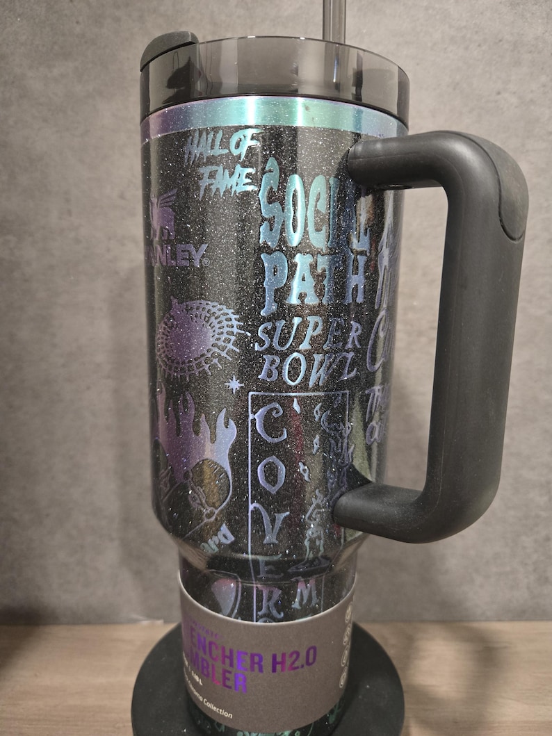 May include: Black glitter tumbler with a black handle and lid. The tumbler features holographic text and graphics, including "Hall of Fame," "Super Bowl," and "Social Path." The tumbler has a straw and is labeled "Quencher H2.0 Tumbler."