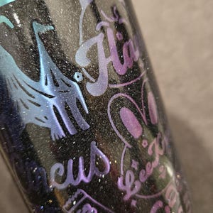 May include: A black glitter tumbler with iridescent blue and purple details. The tumbler has etched designs including a cat, bunny, and the word "H&auml;". Other etched words include "Beez K", "Circus", and "Sracin".