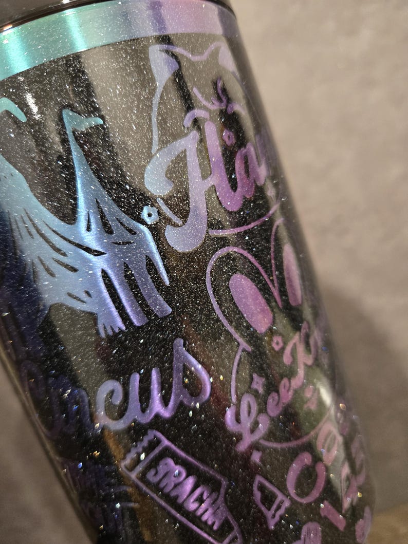 May include: A black glitter tumbler with iridescent blue and purple details. The tumbler has etched designs including a cat, bunny, and the word "H&auml;". Other etched words include "Beez K", "Circus", and "Sracin".