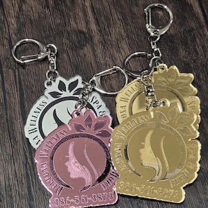 Op de afbeelding: Sleutelhangers met een cirkelvormig ontwerp met een profielsilhouet en de tekst "Deltona Wellness Spa & More" en een telefoonnummer. De sleutelhangers zijn in zilver, roségoud en goud, bevestigd aan zilveren sleutelringen.