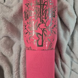 May include: Pink stainless steel tumbler with a silver design of a cross and flowers. The text "worthy" is written on the tumbler. The bottom of the tumbler has the text "The beauty of a woman is her purpose" and a graphic of an arrow.