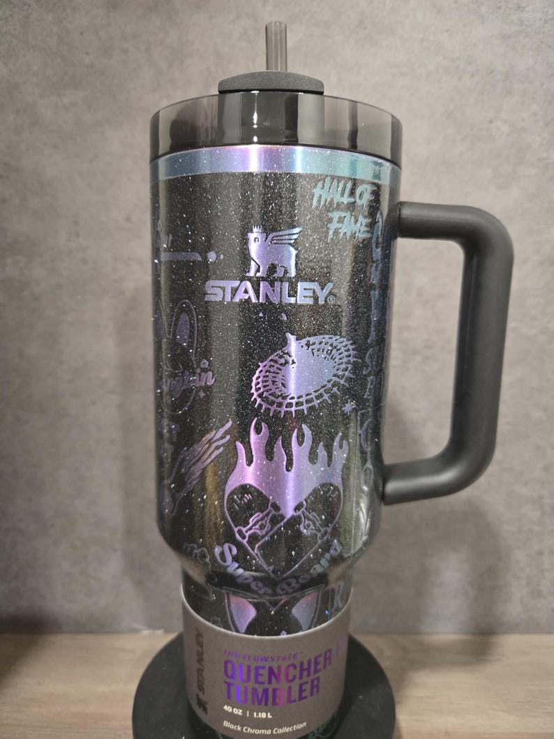 May include: Black Stanley Quencher Tumbler with a handle, featuring a glittery finish and various graphics. The tumbler has a black lid and handle, with the text "HALL OF FAME" and "STANLEY" visible. The tumbler holds 1.18L.