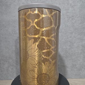 May include: A gold-colored tumbler with a sunflower design and a gray lid. The tumbler has a glittery finish and a giraffe-like pattern in gold. The sunflower design is etched into the tumbler's surface.