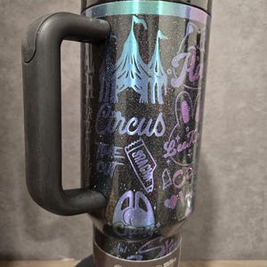 May include: Black glitter travel mug with a handle, featuring a circus-themed design. The design includes circus tents, text that says "Circus", and other circus-related graphics in shades of blue, purple, and pink. The mug has a black lid and a straw.