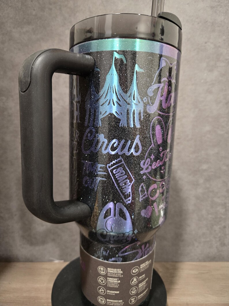May include: Black glitter travel mug with a handle, featuring a circus-themed design. The design includes circus tents, text that says "Circus", and other circus-related graphics in shades of blue, purple, and pink. The mug has a black lid and a straw.