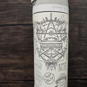 Op de afbeelding: Een witte Owala-waterfles met een grijze dop en een gedetailleerde grijze grafische print. Het ontwerp bevat een auto, een pentagram, een Kansas-kentekenplaat met KAZ 2Y5 en tekst zoals GOT SALT?, CARRY ON MY WAYWARD SON en SALVAGE.