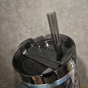May include: Black travel mug with a black lid and a black straw. The mug features a design with purple and white accents. The mug is designed to keep drinks hot or cold.