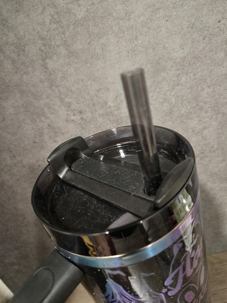 May include: Black travel mug with a black lid and a black straw. The mug features a design with purple and white accents. The mug is designed to keep drinks hot or cold.