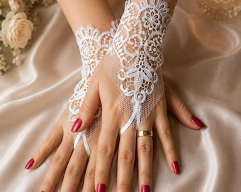 Puede incluir: Un par de guantes de encaje blanco sin dedos con un intrincado patrón floral y una base de malla delicada. Los guantes se extienden desde la muñeca hasta el dorso de la mano, asegurados por un fino lazo alrededor del dedo medio, con bordados.