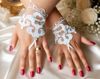 Puede incluir: Un par de guantes de encaje blanco sin dedos con un intrincado patrón floral bordado y un delicado borde festoneado. Los guantes están diseñados para cubrir el dorso de la mano e incluyen una fina lazada blanca que se sujeta al dedo medio.