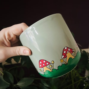 May include: A light green ceramic mug with a white interior and a hand-painted design of red and yellow mushrooms on a green background.