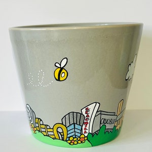 May include: A grey ceramic planter with a hand-painted design of a city skyline, a bee, and a cloud. The planter has a green base and features the words "Crucible" and "Beadmall".