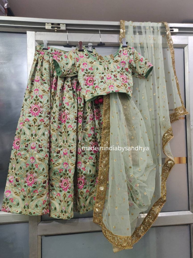 May include: A light green lehenga with floral embroidery and sequins. The lehenga includes a matching embroidered blouse and a sheer dupatta with a gold border.