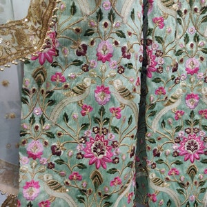 May include: A green fabric with intricate embroidery featuring pink and white flowers, birds, and gold accents. The fabric has a traditional Indian design.