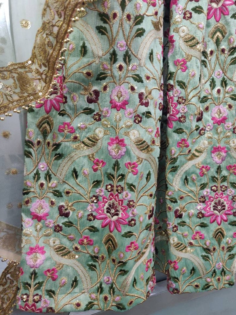 May include: A green fabric with intricate embroidery featuring pink and white flowers, birds, and gold accents. The fabric has a traditional Indian design.