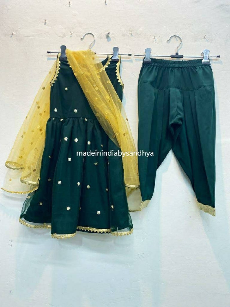 Puede incluir: Un conjunto verde y dorado para un ni&ntilde;o, que consta de un vestido sin mangas con un dupatta de red dorada y pantalones a juego. El vestido tiene lentejuelas doradas y un ribete dorado. El texto "madeinindiabysandhya" es visible en el vestido.