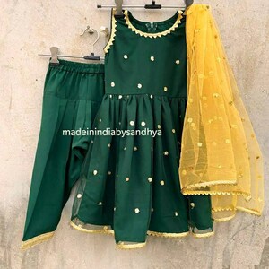 Puede incluir: Un conjunto para ni&ntilde;a en verde y dorado, con una blusa sin mangas, pantalones y una dupatta transparente. La blusa y los pantalones tienen un patr&oacute;n de puntos y ribetes dorados. La dupatta es amarilla con puntos dorados y ribetes dorados.
