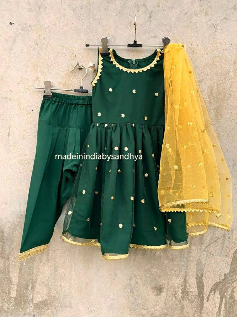 Puede incluir: Un conjunto para ni&ntilde;a en verde y dorado, con una blusa sin mangas, pantalones y una dupatta transparente. La blusa y los pantalones tienen un patr&oacute;n de puntos y ribetes dorados. La dupatta es amarilla con puntos dorados y ribetes dorados.