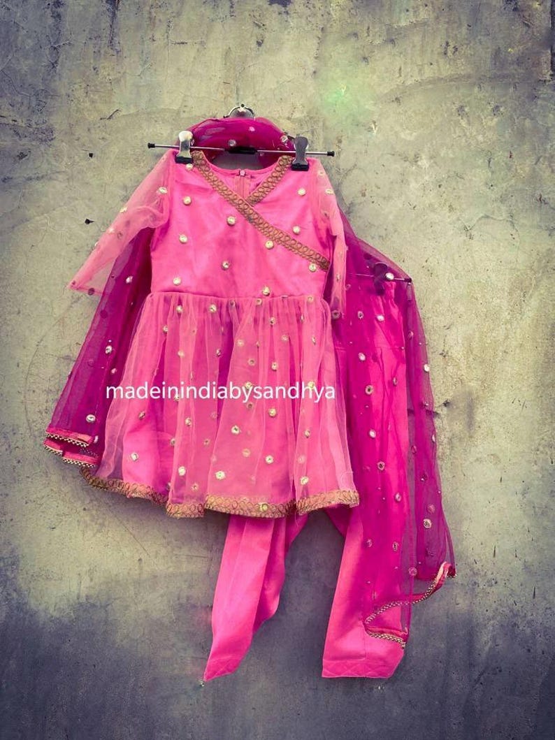 Puede incluir: Un conjunto de ni&ntilde;a rosa y dorado con una blusa de manga larga, una falda y una dupatta a juego. El conjunto est&aacute; decorado con peque&ntilde;as lentejuelas redondas de espejo. El texto "madeinindia bysandhya" es visible en la falda.