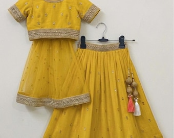 May include: A yellow two-piece outfit, including a short-sleeved top and a skirt, hanging on a rack. The top and skirt are adorned with gold embroidery and small embellishments. The skirt has a sheer overlay.